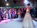 Una mujer vestida con un vestido blanco y corona se encuentra en el centro de una escena de concurso de belleza, rodeada por fotógrafos y asistentes. La escena está iluminada con luces de estudio, destacando a la mujer y sus detalles.