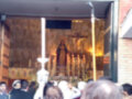 Una procesión religiosa con una imagen de la Virgen María y un crucifijo en el centro, rodeada por fieles que llevan velas y mantas blancas. La escena se desarrolla en un ambiente religioso, con una estructura que parece ser una iglesia o un templo.