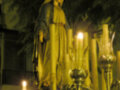 Una estatua de la Virgen María iluminada por velas encendidas, en un ambiente nocturno con sombras y luces difuminadas. La estatua parece ser de madera o metal, con detalles finos y una corona en la cabeza. A su alrededor se pueden ver varios candelabros y velas encendidas, creando una atmósfera de reverencia y devoción.