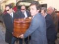 Un grupo de hombres en trajes azules y corbatas llevan un ataúd de madera roja, mientras una furgoneta funeraria con flores y un edificio de atrás.