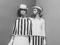 Dos modelos posan con trajes de estilo marinero, compuestos por chaquetas blancas y pantalones cortos con rayas negras. El estilo se asemeja al de los años 60, con sombreros de copa y detalles en blanco.
