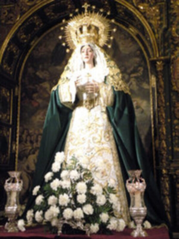Virgen María en iglesia, vestida de blanco con capa verde y corona dorada. Fondo dorado con detalles artísticos. Base de flores blancas y velas en latón.