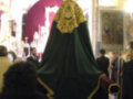 Imagen de una procesión religiosa en un templo, con una imagen de la Virgen María envuelta en un manto verde y dorado. La imagen se encuentra en el centro de la imagen, rodeada por personas que parecen participar en la procesión. En el fondo se pueden ver vitrales y una araña de luz que ilumina la escena.