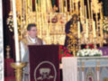 Imagen de una iglesia con un altar central, adornado con velas y flores. En el fondo, una imagen religiosa esculpida en madera. A la izquierda del altar, un sacerdote con una saya y un hábito.