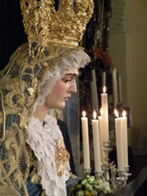 Estatua religiosa con corona dorada y velas encendidas en un ambiente de iglesia.
