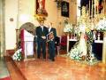 Dos hombres en traje negro se encuentran frente a una imagen religiosa en un altar. La imagen está decorada con flores y adornos, y hay una cruz visible en el suelo. La atmósfera es solemnemente reverente, típica de un lugar de culto.