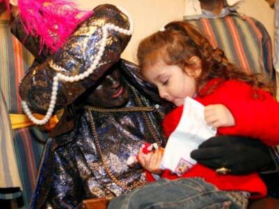 Un hombre disfrazado con un sombrero de plumas y una chaqueta brillante está sentado, mientras una niña con una chaqueta roja le ofrece un regalo.