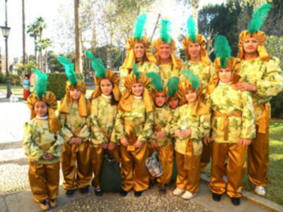 Un grupo de niños disfrazados con trajes verdes y amarillos, vestidos como nativos americanos. Ellos llevan sombreros con plumas y pantalones largos de color marrón. La escena está en un parque con árboles y una calle de fondo.