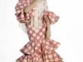 Una mujer vestida con un traje de flamenca rosa con puntos blancos, falda con volantes y mangas largas con flecos. La imagen también muestra un tocado floral en su cabeza.