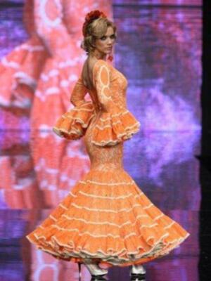 Una modelo presenta un vestido de flamenco naranja con volantes en la falda y mangas con volantes en un desfile de moda.