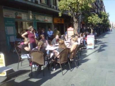 Un grupo de personas disfruta de una comida al aire libre en un café. La calle está llena de gente sentada a mesas con sillas, y hay una fuerte presencia de sombrillas para protegerse del sol. La imagen muestra un ambiente animado y relajado, típico de una zona comercial o turística.