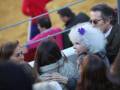 Una niña con una peluca blanca y un lazo morado en el cabello, abraza a una mujer mayor con una chaqueta gris. La escena parece ser en un evento público, con personas de diferentes edades y estilos vestidos.