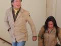 Una persona con una chaqueta beige y camisa amarilla grita mientras otra se tambalea, ambos en un entorno interior con paredes blancas.