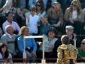 Un torero con un traje de luces y una capa roja se encuentra en el centro de la imagen, frente a un público que incluye adultos y niños. La gente está sentada en gradas y parece estar observando el evento, con algunos de ellos mirando al torero. La escena se desarrolla en un entorno que parece ser una plaza de toros, con el suelo visible y la estructura típica de las gradas.