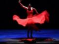 Una bailarina en un vestido rojo volador se eleva sobre la escena, iluminada por una luz que resalta su movimiento y el volumen de su vestido.