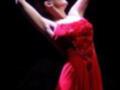 Una bailarina en un vestido rojo brillante, con las manos levantadas en una pose de ballet. La escena es dramática y intensa, con un fondo oscuro que resalta su figura.