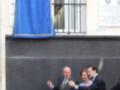 Una bandera azul ondea en el aire, mientras tres personas en trajes de negocios sostienen un cinturón rojo. La imagen se centra en la celebración de una inauguración o ceremonia, con un edificio al fondo.
