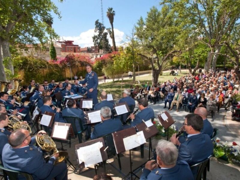 Concierto militar en un parque, con militares tocando instrumentos musicales y público asistente. El ambiente es formal y el clima parece ser de verano con árboles y flores en el fondo.