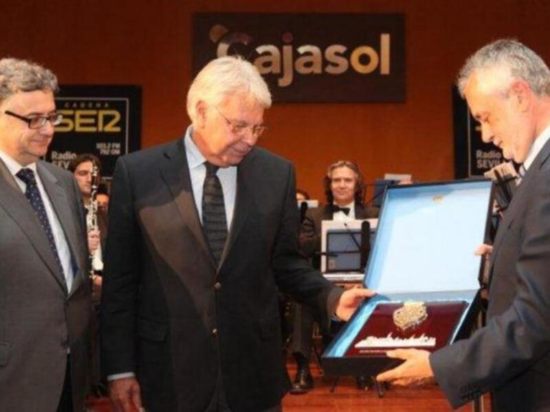 Un hombre en traje negro y corbata azul está entregando una medalla dorada a otro hombre en traje negro y corbata blanca. El fondo muestra un cartel con la palabra 'Cajasol' y un logo de radio.