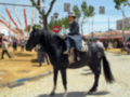 Un hombre monta un caballo negro en una feria. El fondo muestra estructuras de color rojo y amarillo, con un cielo azul claro.