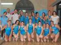 Equipo femenino de natación en una piscina, todos con trajes azules y sonrientes.