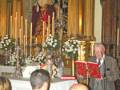 Una iglesia con un altar dorado, una imagen de Cristo y flores blancas. Un hombre en traje gris habla a dos personas sentadas, uno de ellos con una bufanda roja.