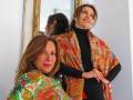 Dos mujeres posan con bufandas de colores vibrantes frente a un espejo. La escena parece ser en un ambiente elegante, posiblemente una boutique o estudio de moda.