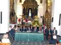 Una reunión formal en una iglesia con un altar central, sacerdotes y personas sentadas alrededor de una mesa.