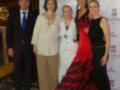 Una familia posa juntos en un evento formal, con una mujer vestida de rojo y otra de negro. Detalles como el fondo con logotipos de hotel y la presencia de un hombre en traje azul destacan.