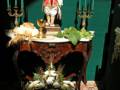 Una escena festiva con un joven en traje colonial sentado en una mesa de madera tallada. La mesa está adornada con flores blancas y un ramo de hojas verdes, junto a velas azules en candelabros. La pared detrás es verde y el suelo tiene un patrón de cuadros.