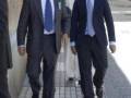 Dos hombres en trajes de negocios caminan por una calle. La imagen muestra a dos personas vestidas formalmente, uno con un traje azul y corbata azul, y la otra con un traje negro. La mujer a su lado lleva un vestido blanco largo y zapatos de tacón. En el fondo, se pueden ver edificios y una calle con gente caminando.