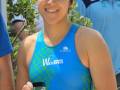 Una mujer con una camiseta azul y verde, sonriendo mientras sostiene un trofeo. Detrás de ella, hay dos hombres que también están sonriendo. La escena parece ser al aire libre con vegetación en el fondo.