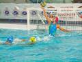 Juego de waterpolo en una piscina con jugadores usando cascos azules y amarillos, uno de ellos lanzando la pelota hacia el arco marcado con "Concepción" y "Ciudad Línea".