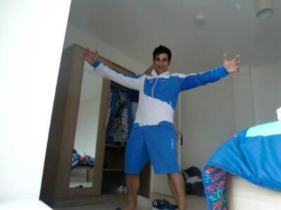 Un hombre en ropa deportiva azul y blanca se muestra con los brazos extendidos, frente a un espejo grande. Detrás de él se ve una estantería con ropa y un armario abierto.