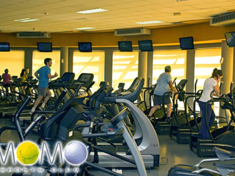 Gente haciendo ejercicio en una sala de fitness con pantallas de televisión en el techo y una marca de deportes en la pared.