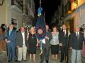 Celebración de una ceremonia en una calle histórica de noche, con personas en trajes y una bandera central.