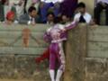Un torero en traje de cuero púrpura y blanco se inclina sobre un muro mientras sostiene una capa roja. La imagen muestra a varios espectadores observando la escena desde arriba, en un ambiente de toros.