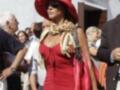 Una mujer viste un vestido rojo con un sombrero y una bufanda, camina entre personas en un entorno urbano.
