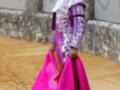Un torero en traje tradicional de color púrpura y blanco, con un faldón rosa brillante, en una plaza de toros.