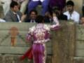 Un torero en traje morado y blanco con una capa roja, preparándose para un combate. En el fondo, espectadores observan la escena con interés.