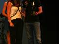 Una mujer y un hombre están en una escena de premiación. La mujer, vestida con un top blanco y pantalones negros, está sonriendo mientras el hombre, que lleva una camiseta negra y pantalones vaqueros, sostiene un trofeo. Ambos parecen estar en una celebración o evento formal.