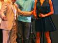 Una mujer en un vestido azul sostiene un trofeo mientras está rodeada por tres personas: una mujer mayor en traje, un hombre joven con camisa azul y otra mujer en el fondo. La escena parece ser una ceremonia o evento formal con un fondo azul y una iluminación suave.