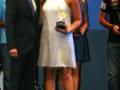 Una mujer en un vestido blanco con flecos sostiene un premio mientras está rodeada por personas en una escena formal.