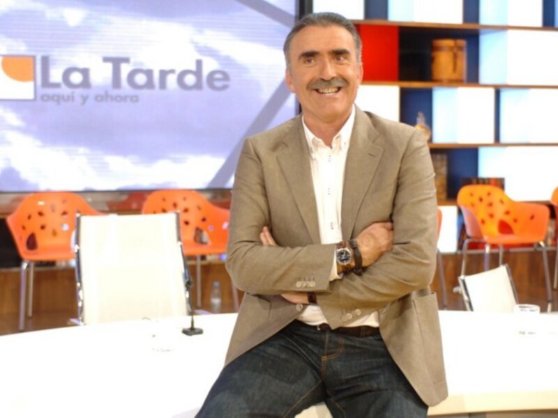Un hombre con una chaqueta beige y camisa blanca, posando con las manos cruzadas en un estudio de televisión con una pantalla de 'La Tarde' en el fondo.