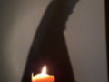 La sombra de una figura se proyecta sobre un fondo claro, con la parte superior del cuerpo y el brazo extendido hacia arriba. En el frente de esta sombra, se observa una vela encendida que emite un resplandor rojizo.
