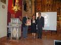 A tres personas en un entorno religioso, posiblemente una iglesia. La persona central sostiene un premio o reconocimiento mientras las otras dos observan con interés. En el fondo, se puede ver una pantalla proyectando un dibujo y estatuas religiosas. La decoración y el ambiente sugieren una ceremonia o evento de reconocimiento en un lugar de culto.