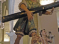 Estatua religiosa de un sacerdote con un cetro y una corona, rodeada por flores rojas.
