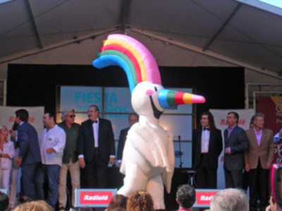 Una figura inflable de un unicornio con una lluvia de arcoíris en la cabeza se encuentra entre un grupo de personas en un escenario con una pancarta que dice "Fiesta". La iluminación es brillante y la atmósfera parece ser de una celebración o evento.