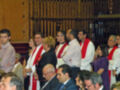 Una imagen de una iglesia donde un grupo de personas, probablemente sacerdotes o seminaristas, están vestidos con vestiduras blancas y rojas. La imagen muestra a varios individuos sentados en filas, algunos con gafas y otros con cabello corto. La decoración interior de la iglesia es visible, incluyendo una estructura de madera y un techo con detalles artísticos.