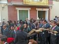 Una procesión religiosa en un entorno urbano, con personas vestidas formalmente y una figura central cargando una cruz.